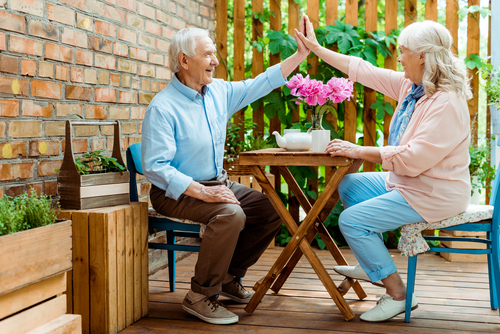 cheerful senior couple giving high five while sitting near pink flowers on terrace 