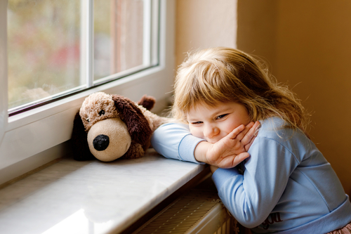 Upset toddler girl sitting by window and looking out. Sad crying child feeling unhappy. Lockdown and isolation concept during corona virus pandemic time. Lonely preschool kid.