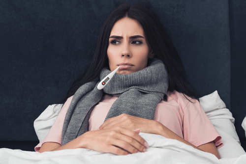 sick young woman measuring temperature with mouth thermometer while lying in bed