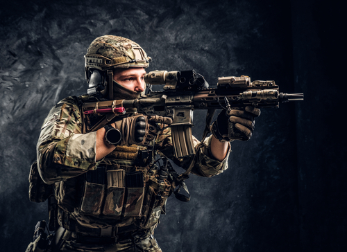 Close-up studio photo against a dark wall. The elite unit, special forces soldier in camouflage uniform holding an assault rifle with a laser sight and aims at the targe